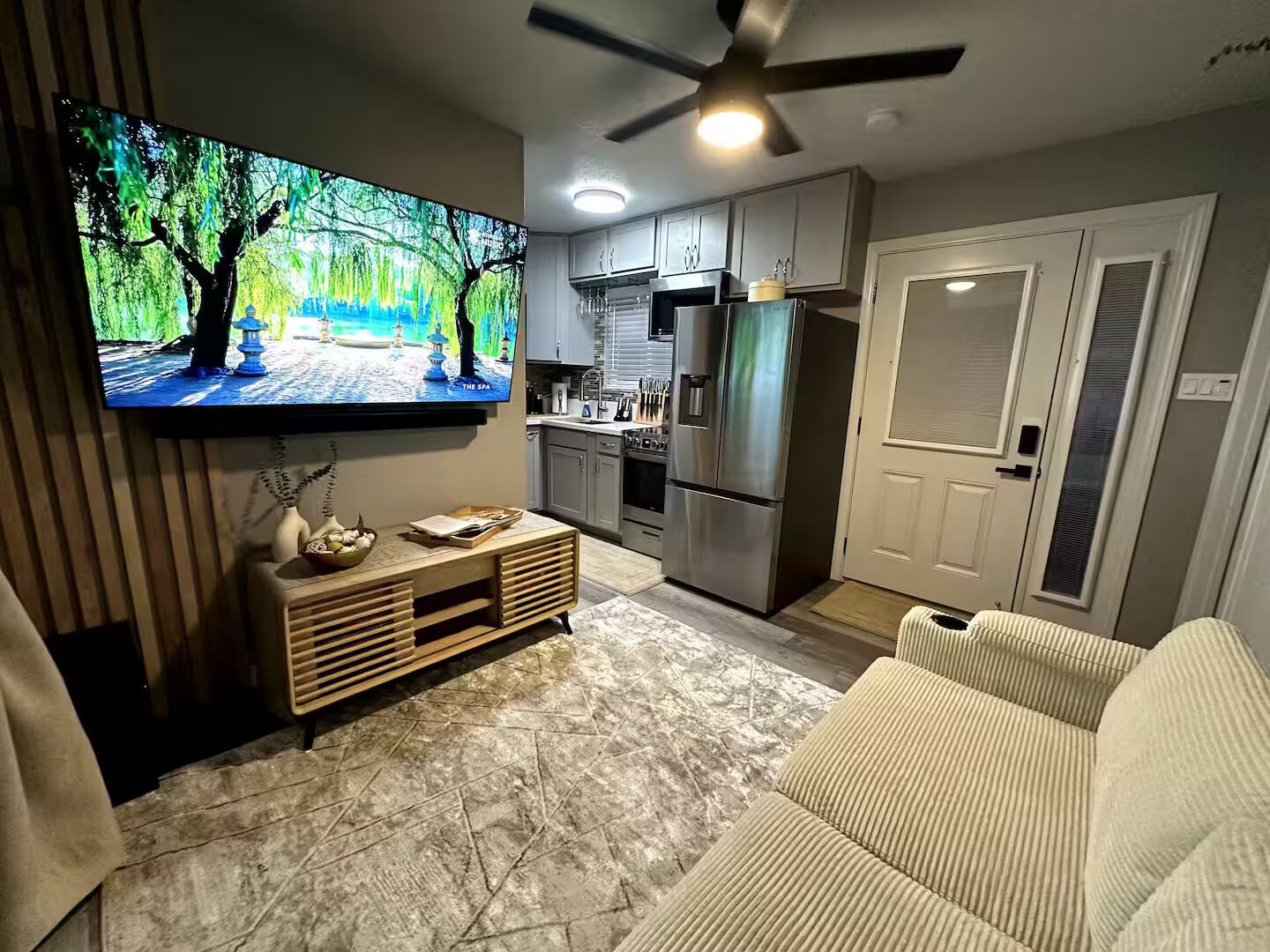Una cocina moderna con horno empotrado, microondas y refrigerador. Hay un armario de madera con un jarrón y una bandeja de fruta. Un televisor está montado en la pared sobre el armario.