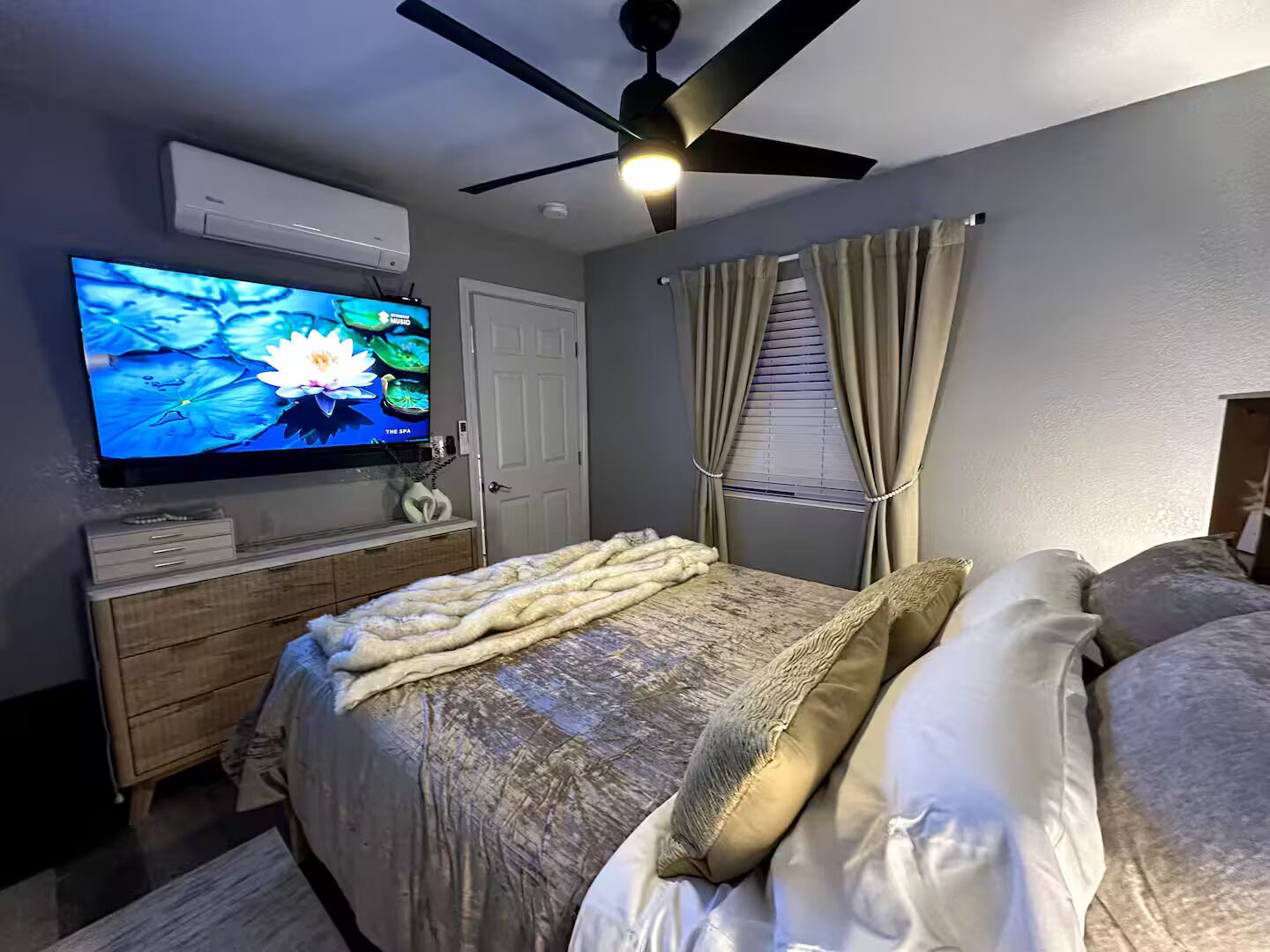 Une chambre à coucher avec un lit, un téléviseur fixé au mur, un ventilateur de plafond et une fenêtre avec des rideaux.