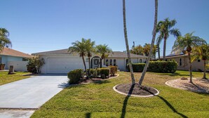 A residential property with a single-story house, a driveway, and a well-maintained lawn with palm trees and shrubs. - Coastal Coral Paradise with Heated Pool (Cape Coral)