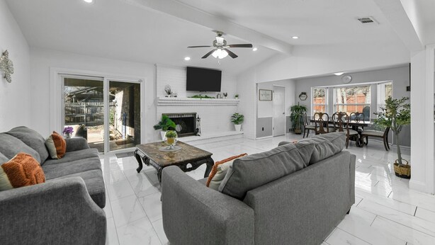 A spacious living area with a grey sofa set, a fireplace, a dining area with a table and chairs, and a ceiling fan. - Diamond Waters near DFW & Stadium with Private Pool (Fort Worth)