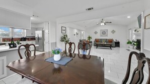 A spacious living area with a dining table, chairs, and a sofa. There is a kitchen area with white cabinets and a refrigerator. - Diamond Waters near DFW & Stadium with Private Pool (Fort Worth)