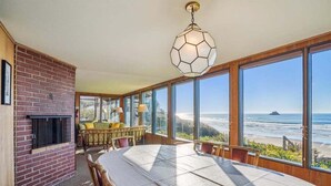 Comedor con una mesa grande, chimenea de ladrillo y ventanas amplias con vistas al océano.