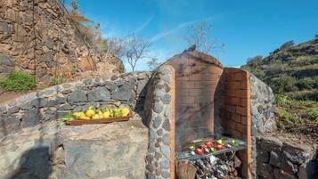 An outdoor stone barbecue grill with a stone wall and a wooden tray of vegetables.