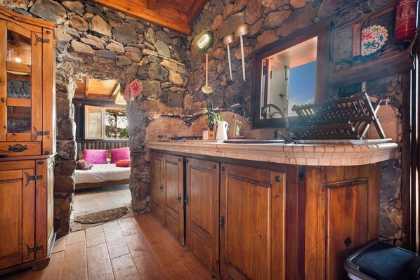 A rustic kitchen with stone walls, wooden cabinets, and a cozy seating area with a pink pillow. - House Finca Relax Cercadillo (Santa Ursula)