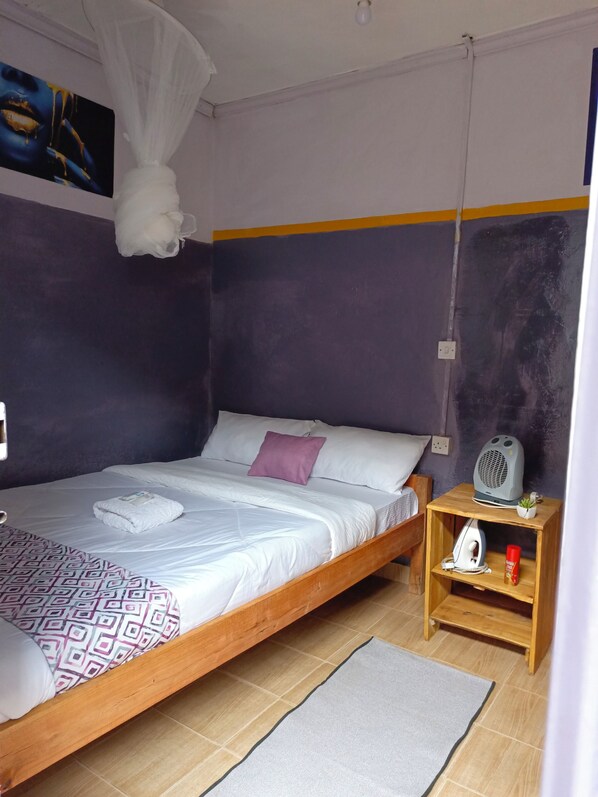 A single bed with a patterned blanket, a bedside table with a heater, and a small plant. - Wrech House 1989 - Sagana (Sagana)