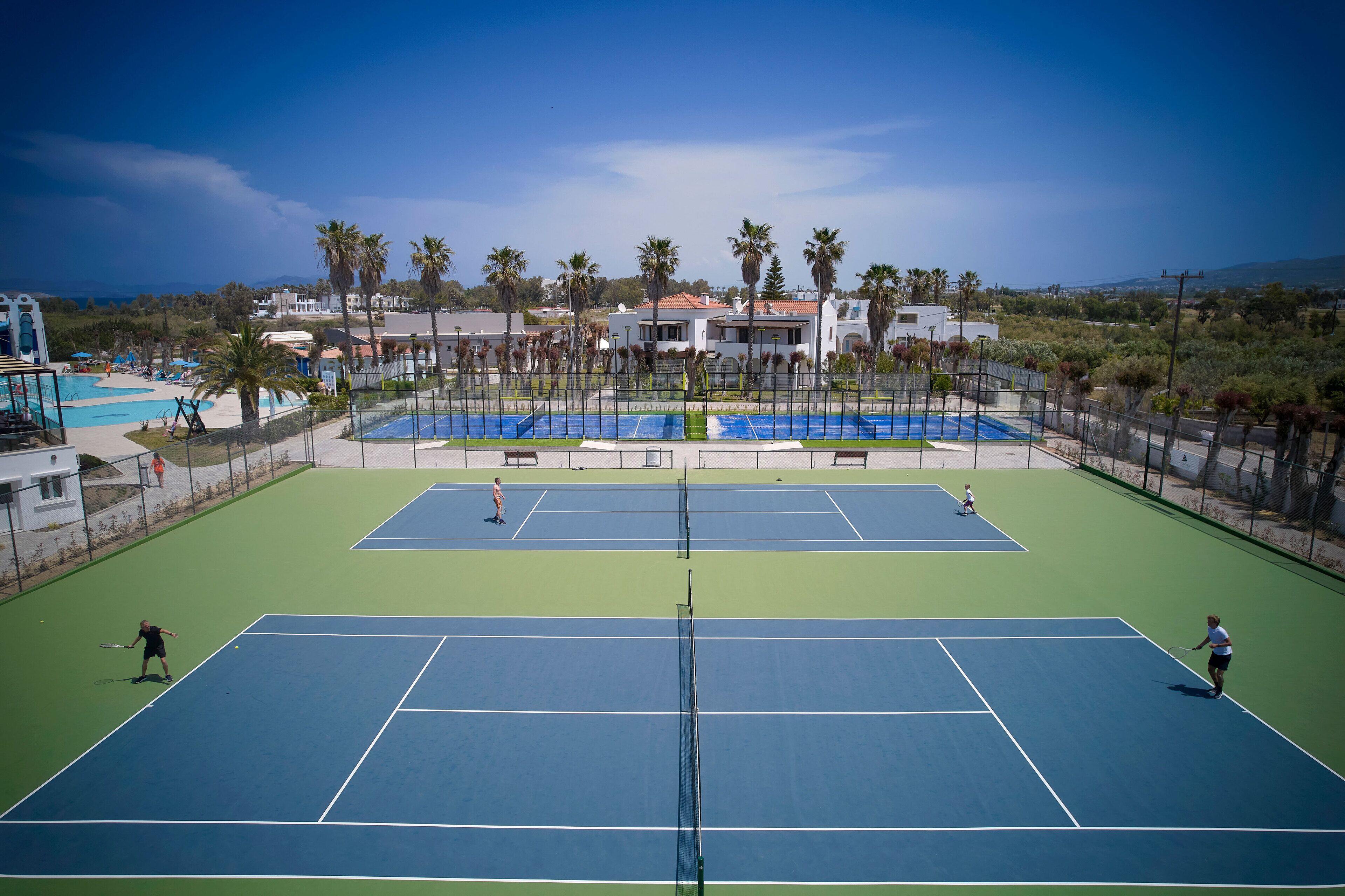 An outdoor tennis court with multiple courts, surrounded by palm trees and buildings.