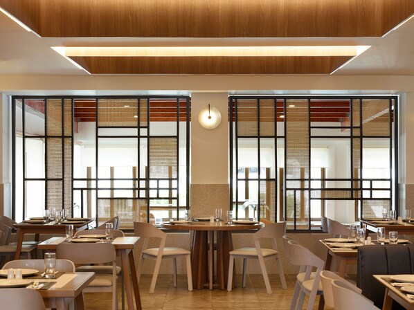 A modern restaurant with wooden tables and chairs, large windows with decorative screens, and a warm ceiling light. - Atlantica Marmari Beach (Kos)