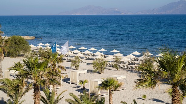 A beach with rows of sun loungers and umbrellas, palm trees, and a view of the sea and distant mountains. - Atlantica Marmari Beach (Kos)