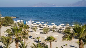 A beach with rows of sun loungers and umbrellas, palm trees, and a view of the sea and distant mountains. - Atlantica Marmari Beach (Kos)