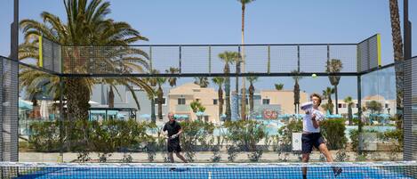 A tennis court with a net, palm trees, and a pool in the background.