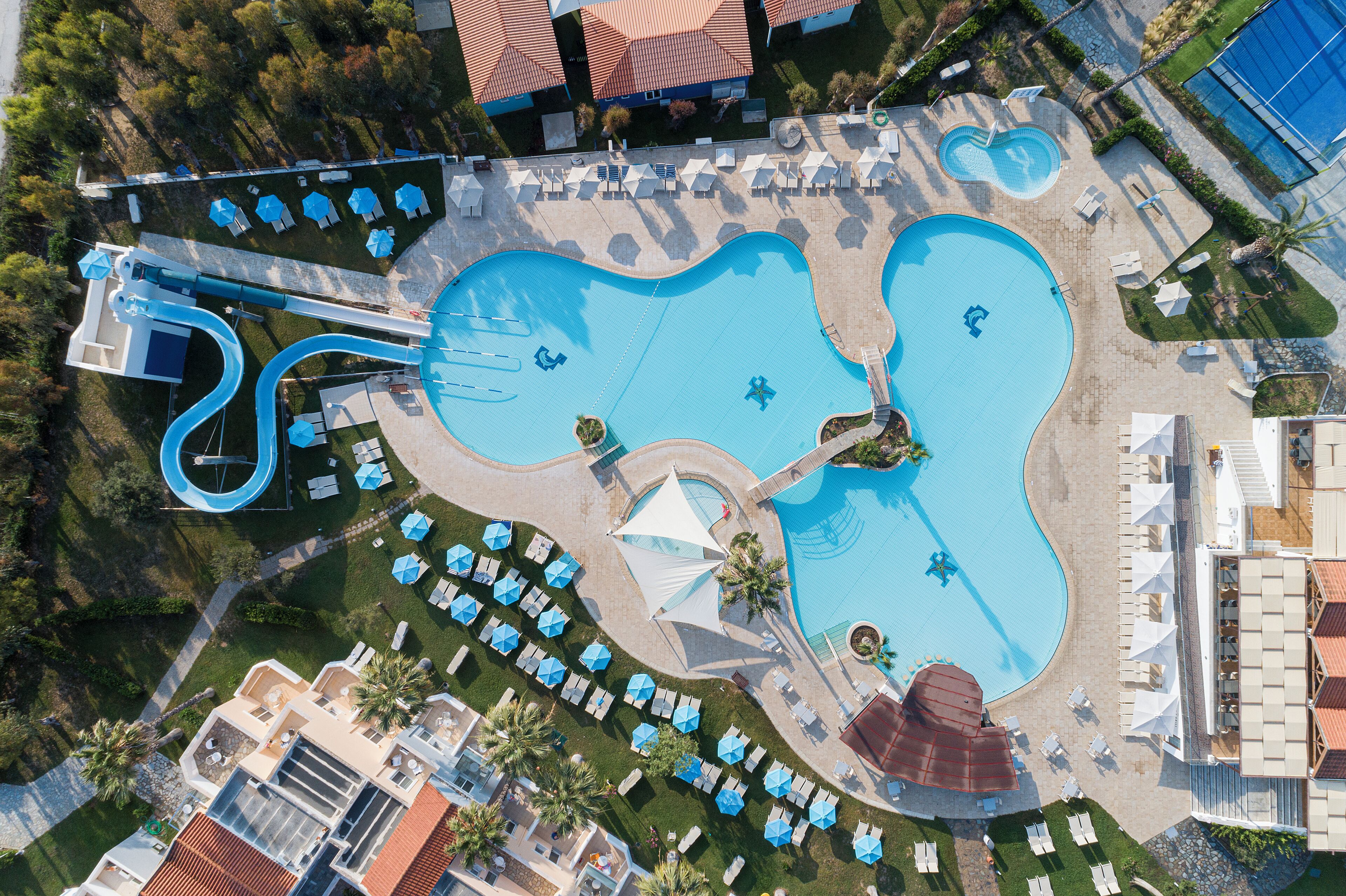 Luftaufnahme eines Resorts mit großem Pool, mehreren Liegebereichen und einer Wasserrutsche.