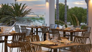 A restaurant with tables set for dining, featuring a view of palm trees and a sunset. - Atlantica Marmari Beach (Kos)