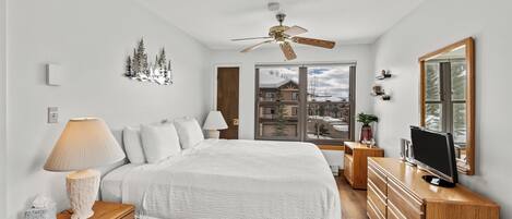 Un dormitorio con cama, mesitas de noche, televisor, ventilador de techo y ventana con vistas a edificios.