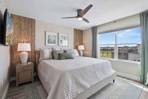Une chambre à coucher comprenant un lit, deux tables de chevet, un ventilateur de plafond, une fenêtre avec vue et des œuvres d’art accrochées au mur.