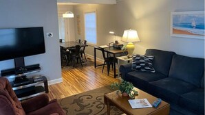 A living room with a flat-screen TV, a sofa, a coffee table, and a dining area with chairs and a table.
