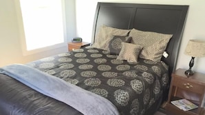 A neatly made bed with a patterned bedspread, a dark headboard, and a bedside table with a lamp and a clock.