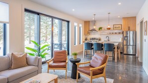 Una sala de estar moderna con un sofá, una mesa de centro y sillones de cuero. Se ve una cocina al fondo con taburetes de bar y un refrigerador.