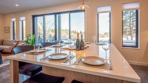 Cocina moderna con una isla grande, abundante luz natural gracias a amplios ventanales, y un comedor con sillas y una mesa dispuesta para una comida.