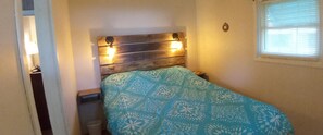 A bedroom with a wooden headboard, a bed with a blue floral bedspread, a small bedside table, and a window with blinds.