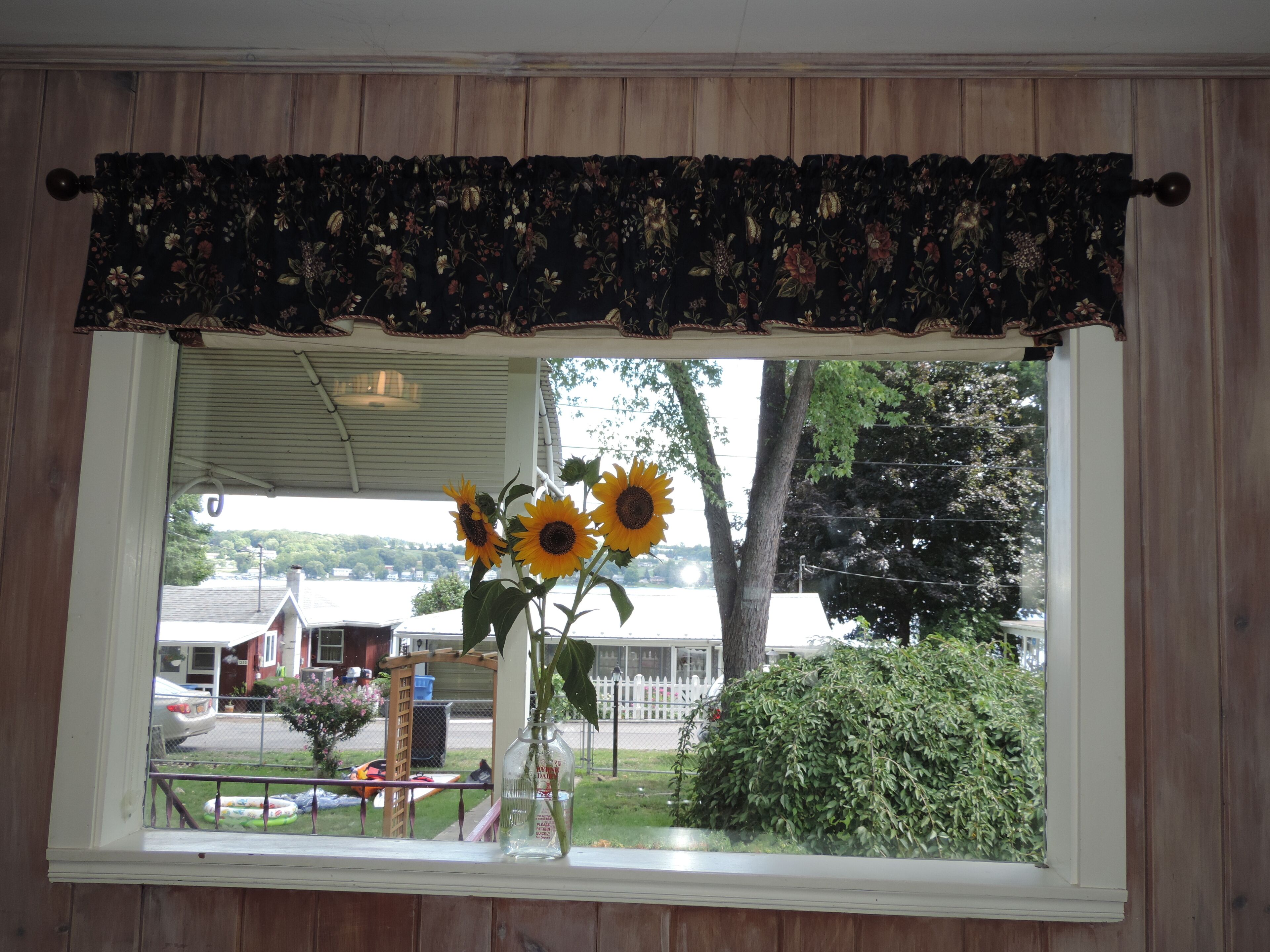 A window with a floral valance, a vase with sunflowers, and a view of a residential area with houses and trees.