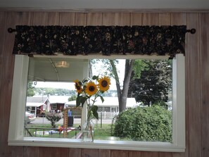 A window with a floral valance, a vase with sunflowers, and a view of a residential area with houses and trees.
