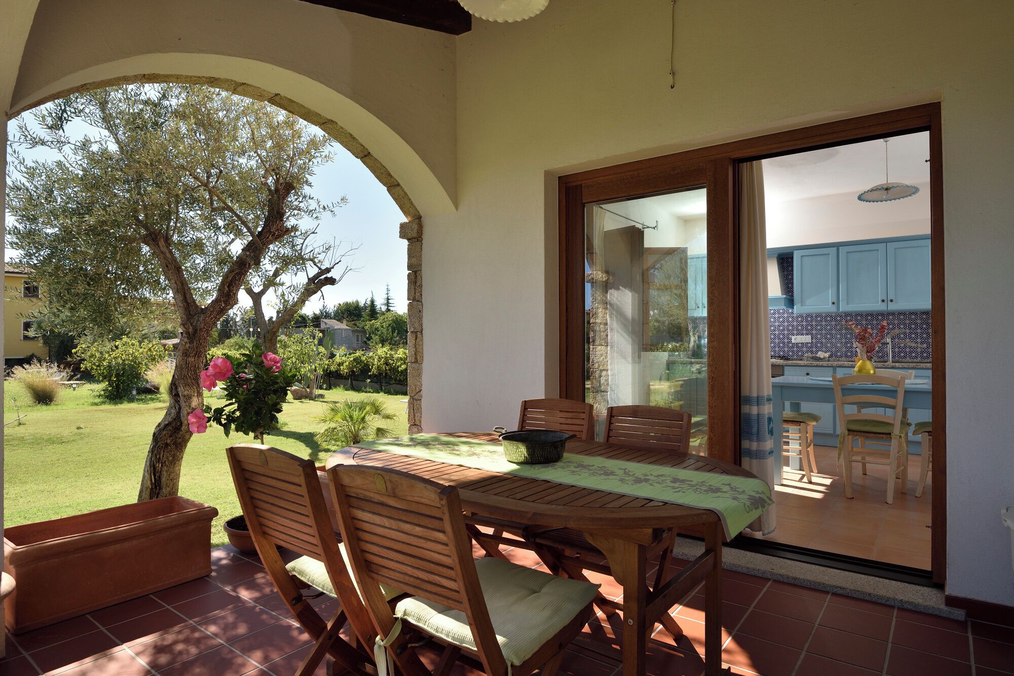 Un patio al aire libre con una mesa y sillas de madera, una planta en maceta y vista a una casa a través de una puerta de cristal.