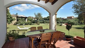 A covered patio with a table and chairs, a view of a garden, and a stone wall in the background. - Villa Menhir! (Sardegna)