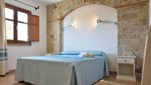 A bedroom with a stone wall, a bed with a blue bedspread, a nightstand, and a wall-mounted lamp. - Villa Menhir! (Sardegna)