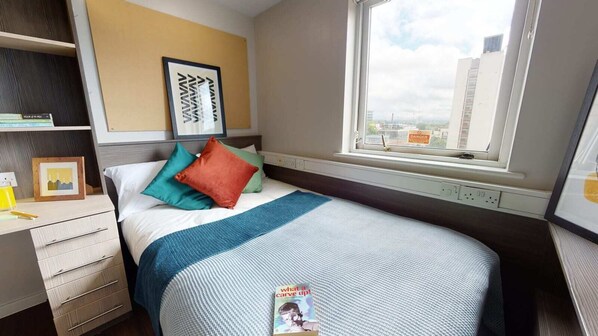 Cozy retreat with a city view, featuring a comfortable bed, a desk for work, and a shelf with personal touches.