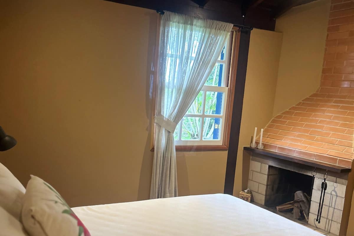 A bedroom with a large bed, a window with curtains, a brick fireplace, and a wooden ceiling.