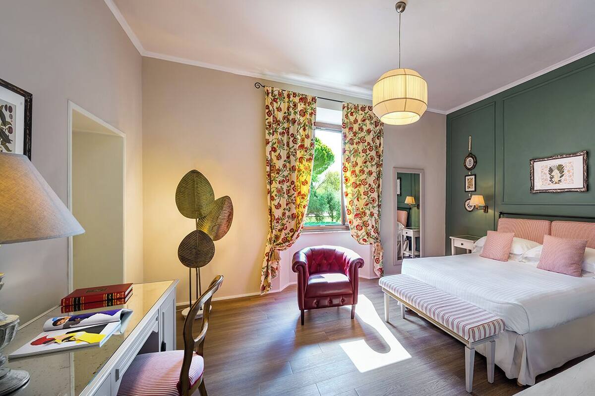 Une chambre à coucher avec un lit, un banc, une chaise, un bureau avec des livres, une lampe et une fenêtre avec des rideaux.