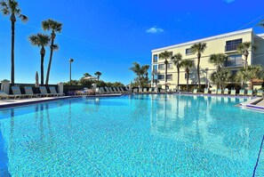 A clear swimming pool with lounge chairs and palm trees, surrounded by a building with balconies.