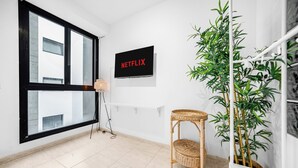 Een kamer met een groot raam, een aan de muur bevestigde televisie waarop 'NETFLIX' staat, een rieten stoel, een pot met bamboe en een witte plank.
