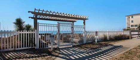 Propiedad privada con acceso mediante una puerta, un porche de madera, cerca blanca y vista despejada de la playa y el océano.