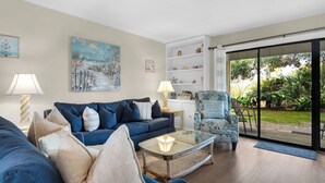 A living room with a blue sofa, a glass coffee table, and a chair with a floral pattern. There is a painting on the wall and a bookshelf with decorative items. - Chateau La Mer II 902 - Pirates Mermaid (Destin)
