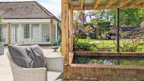 Una veranda coperta con mobili in rattan, uno stagno in mattoni e vista su una casa con ampie finestre e un giardino.
