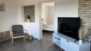 A modern living room with a grey sofa, a TV on a white stand, and a stone wall. - Modern apartment - top location (Witten)