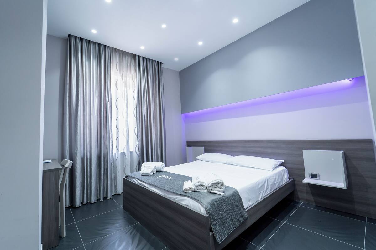 A modern hotel room with a neatly made bed, a bedside table, a wall-mounted charging station, and a window with curtains.