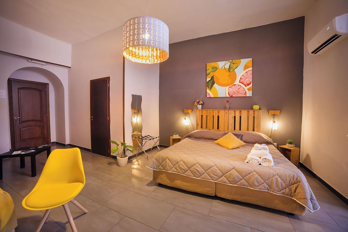 A modern bedroom with a large bed, a bright yellow chair, a hanging light fixture, and a wall-mounted painting.