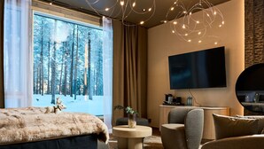 A modern hotel room with a large window offering a snowy forest view, a cozy bed, a flat-screen TV, and a seating area with a round coffee table. - Balder Luxury Lodge & Glass Igloos (Kittila)