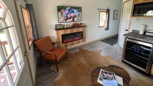A living room with a fireplace, a television mounted above it, a dining area with a kitchen counter, and a door leading outside. - Alpaca Cabin #1 (Audubon)