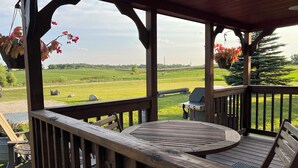 A wooden deck with a round table and chairs, overlooking a green field and a road. - Alpaca Cabin #1 (Audubon)