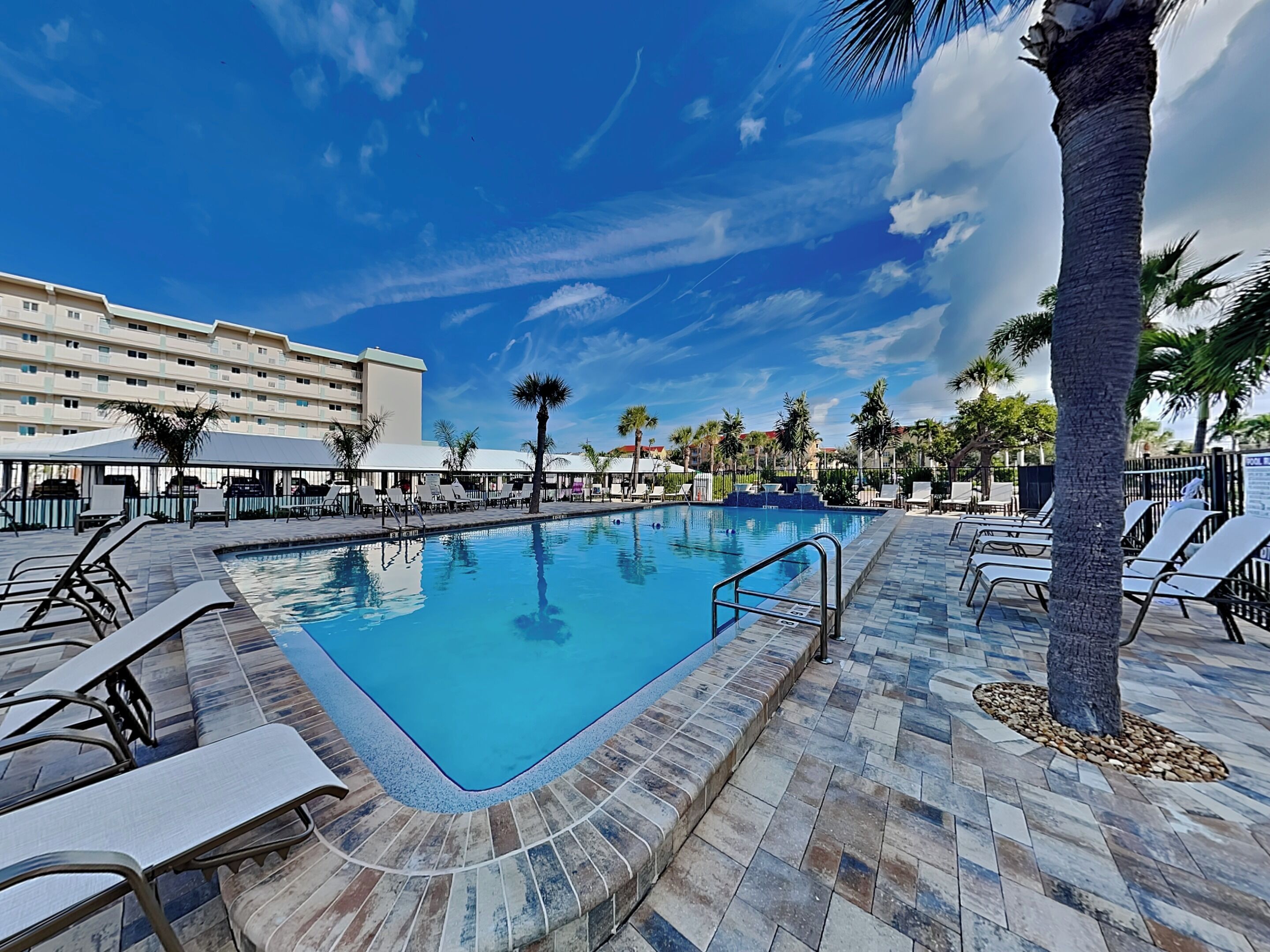 Une piscine entourée de chaises longues et de palmiers, le tout adossé à un bâtiment d’hôtel.