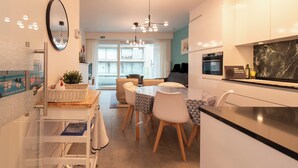 A modern kitchen with a dining area, featuring a central island, white cabinets, and a large window providing natural light. - Newly-built Apartment by the sea (Middelkerke)