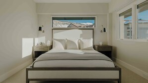 Ein Schlafzimmer mit einem Bett, zwei Nachttischen, einem Fenster mit Blick auf Gebäude und einer Deckenleuchte.
