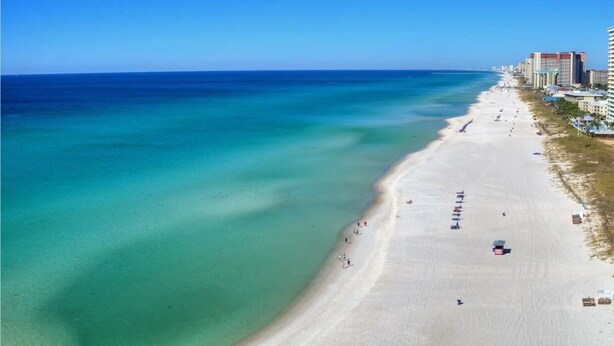 Luchtbeeld van een strand met helder turquoise water, een zandstrand en gebouwen langs de kust.
