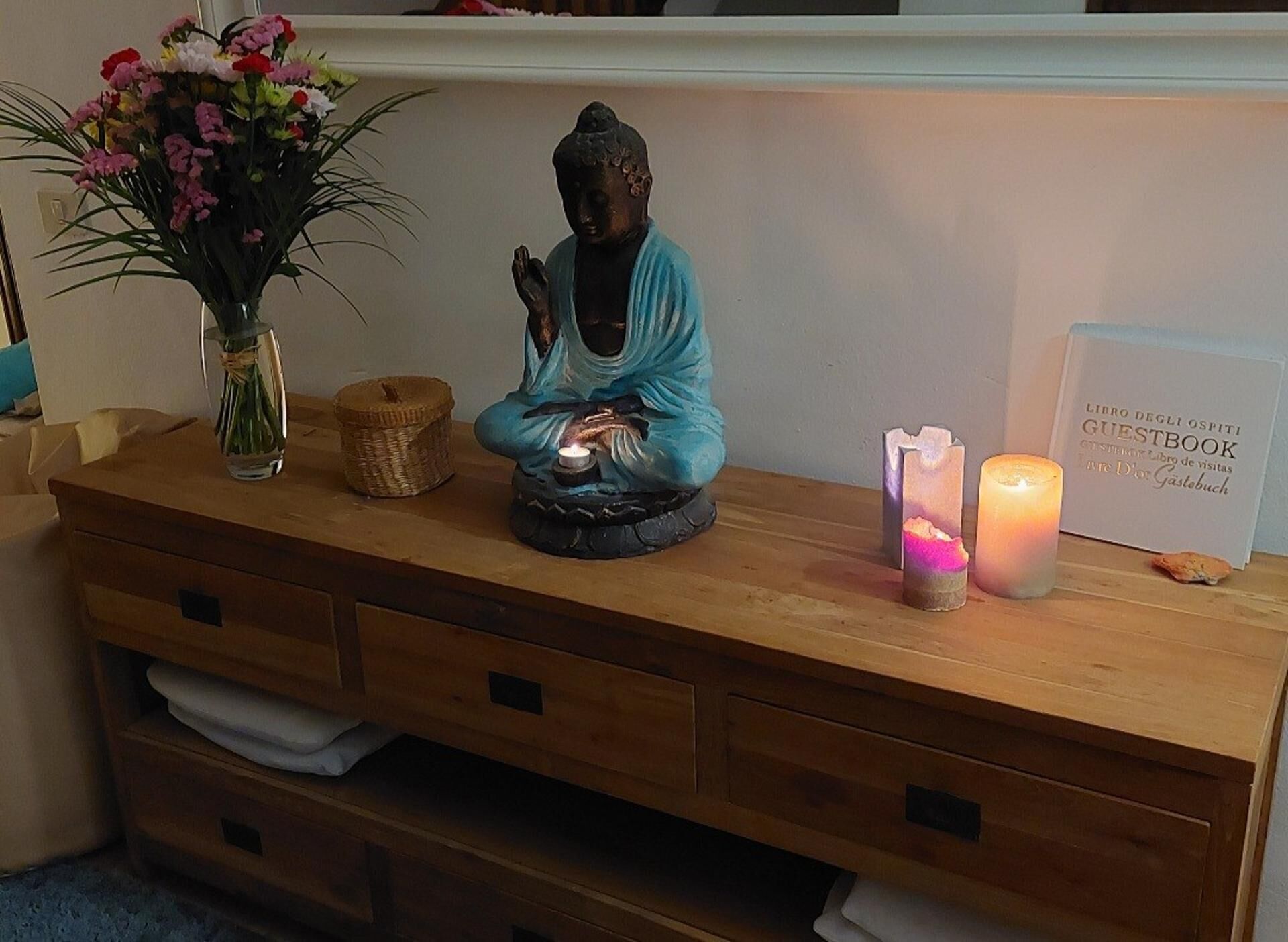 Ein hölzerner Kommode mit Buddha-Statue, Blumen in einer Vase, einer Kerze und einem Gästebuch.
