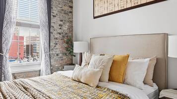 A hotel room with a large bed, a brick wall, a window with curtains, and a framed artwork on the wall.