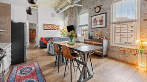 A modern kitchen with a dining area, featuring exposed brick walls, a large window, and a ceiling fan.