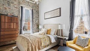 A bedroom with a brick wall, a bed with a yellow and white duvet, a wooden dresser, a nightstand with lamps, a yellow armchair, and a view of buildings through sheer curtains.